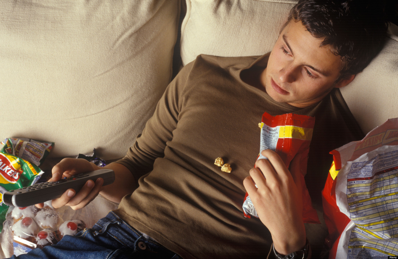 B8NDBN slobbish man lounging on a settee watching television and eating junk food